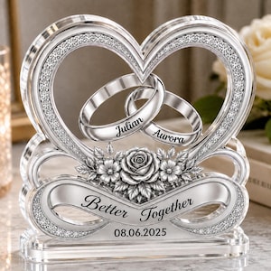 Pode incluir: Um ornamento de acrílico transparente em forma de coração com dois anéis entrelaçados gravados com os nomes Julian e Aurora. O design inclui uma rosa central, uma faixa com o texto Better Together e a data 08.06.2025.