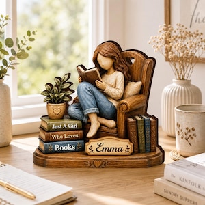 May include: A decorative figurine of a person with brown hair sitting in a wooden armchair reading a book. The base features a stack of three books labeled Just A Girl Who Loves Books and a nameplate reading Emma, with a small potted plant nearby.