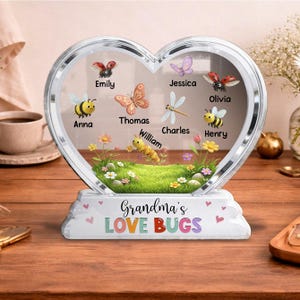 May include: A heart-shaped decorative piece with a clear center, featuring names and insect illustrations. The base reads "Grandma's LOVE BUGS." Includes butterflies, bees, a dragonfly, and a caterpillar. The piece is on a wooden surface.