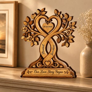 May include: A wooden tree sculpture featuring intertwined trunks forming a heart shape, with engraved names Daniel and Jessica, a heart-shaped tag with the date 05.06.2026, and a base inscribed with the text Our Love Story Began, decorated with leaves and flowers.