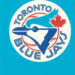Peut inclure: Un cercle blanc avec les mots "Toronto Blue Jays" en police rouge, blanche et bleue. Un geai bleu avec une feuille d'érable rouge est au centre du cercle.