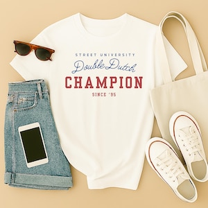 May include: A white t-shirt with the text "Street University Double Dutch Champion Since '95" in blue and red. The shirt is paired with denim shorts, sunglasses, a tote bag, white sneakers, and a smartphone.