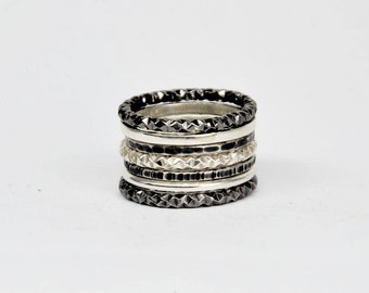May include: A stack of six metal rings featuring alternating textures and finishes. The set includes three dark, oxidised rings with faceted geometric patterns, two smooth polished silver-tone bands, and one band with a textured, hammered surface.