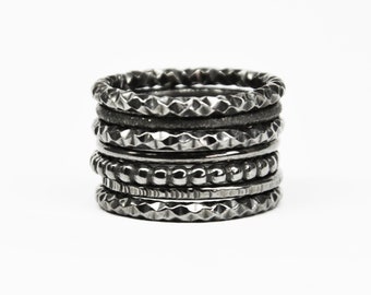 May include: A stack of six dark metallic rings with varying textures. The set includes bands with faceted geometric patterns, a smooth matte band, and a band with a beaded surface, all featuring a consistent dark gunmetal grey finish.
