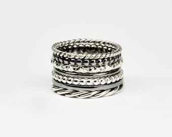May include: A stack of silver and black rings with different textures, including rope, bead, and geometric patterns.