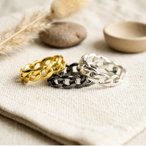 Puede incluir: Tres anillos de eslabones entrelazados dispuestos en grupo. Un anillo tiene un acabado en tono dorado brillante, otro en gris plomo oscuro y otro en tono plateado pulido. Cada anillo presenta un patrón de cadena de bordillo en toda la banda.