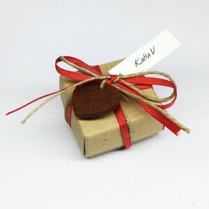 May include: A small, brown gift box tied with red ribbon and twine. A brown felt tag is attached to the ribbon, and a white tag with the text "Katia V" is attached to the twine.