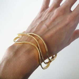 May include: Three gold-toned metal bangles with a wavy design. The bangles are stacked on top of each other on a person's wrist.