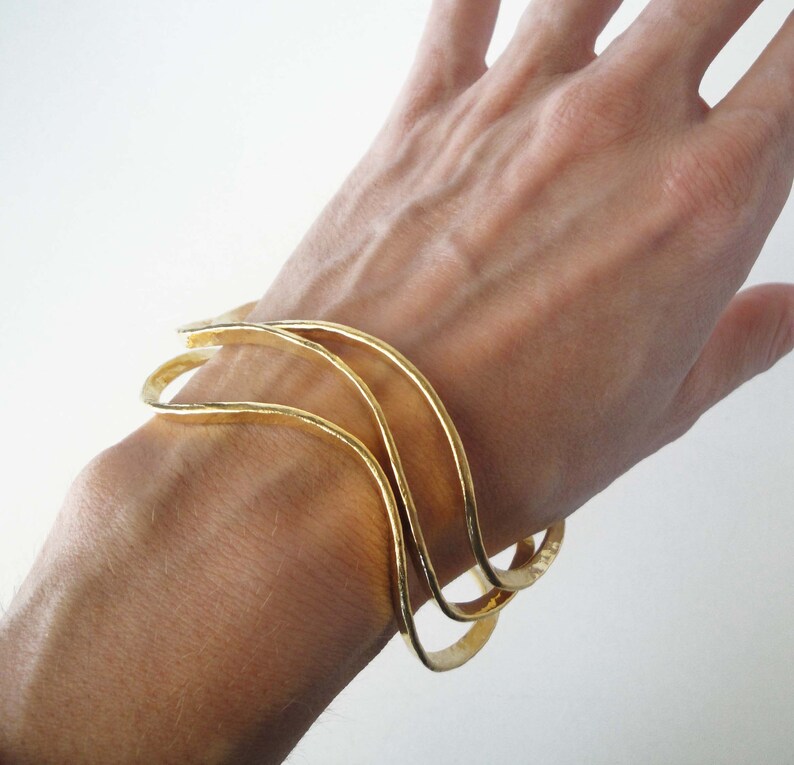 May include: Three gold-toned metal bangles with a wavy design. The bangles are stacked on top of each other on a person's wrist.