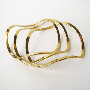 May include: Three gold-toned metal bangles with a wavy, organic shape. The bangles are stacked on top of each other.