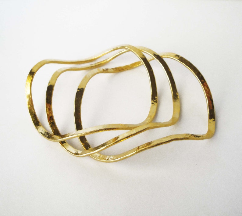 May include: Three gold-toned metal bangles with a wavy, organic shape. The bangles are stacked on top of each other.