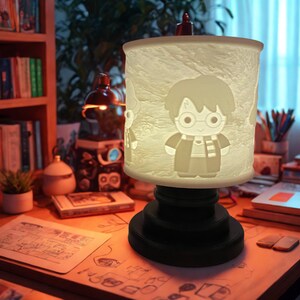 Può includere: Una lampada cilindrica con base nera e paralume bianco con un design a personaggio dei cartoni animati. La lampada è su una scrivania di legno con libri e altri oggetti. La lampada è accesa, emettendo un bagliore caldo.