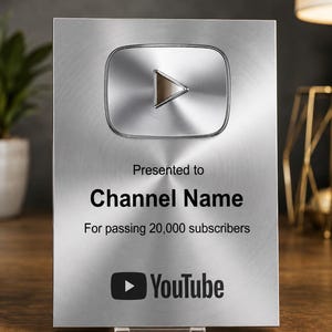 May include: A rectangular silver-toned metal plaque featuring a raised YouTube play button icon at the top. The text reads: Presented to Channel Name, For passing 20,000 subscribers, and includes the YouTube logo at the bottom centre of the surface.