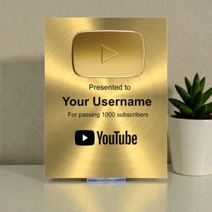 May include: A gold-colored YouTube Play Button award with a brushed metal finish. The award features the YouTube logo and text that reads "Presented to Your Username For passing 1000 subscribers."