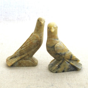 May include: Two carved stone dove figurines. The birds are in a light tan color with darker streaks, and are perched on small bases. The doves are side-by-side, facing different directions, and are set against a neutral background.
