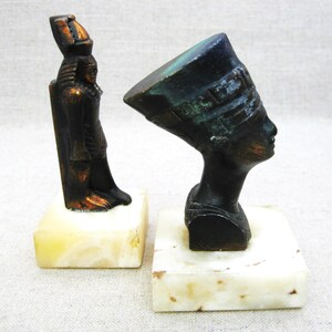 May include: Two small bronze statues of Egyptian figures on marble bases. The statue on the left is of a standing figure with a tall headdress. The statue on the right is of a bust of a woman wearing a headdress.