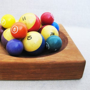 May include: A wooden bowl filled with billiard balls in various colors, including red, yellow, blue, green, and brown. The balls are numbered with black paint.