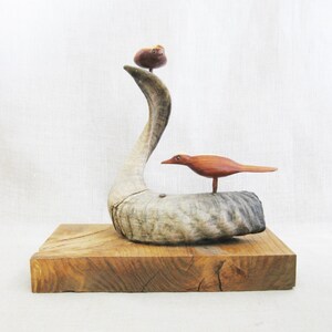 May include: A wooden sculpture of a bird perched on a curved piece of driftwood, with a smaller bird on top. The sculpture is mounted on a wooden base.