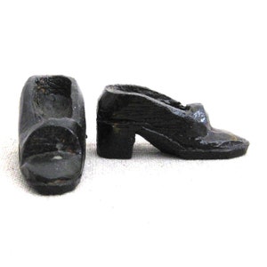 May include: Two small, antique-style, black wooden shoes. One shoe is upright, showing the opening for the foot. The other shoe is on its side, revealing a small heel. The shoes appear to be hand-carved and have a distressed finish.