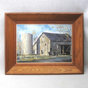 May include: A framed painting of a rural scene featuring a weathered barn and a silo under a blue sky. The barn has a dark roof and is set against a backdrop of trees and a road. The frame is made of wood.