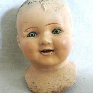 May include: Antique doll head with a painted face, featuring blue eyes, and a slightly open mouth. The doll head has a cracked, aged appearance, with peeling paint on the forehead and neck. The doll head is a vintage collectible.