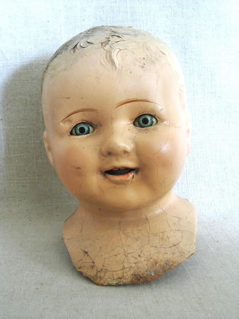 May include: Antique doll head with a painted face, featuring blue eyes, and a slightly open mouth. The doll head has a cracked, aged appearance, with peeling paint on the forehead and neck. The doll head is a vintage collectible.