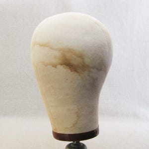 May include: A beige wig head with brown staining, mounted on a dark brown base and a metal stand. The head is oval-shaped and has a smooth surface. The stand is a dark metal color.