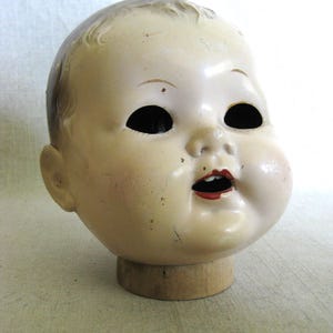 May include: Vintage doll head with a pale complexion, black eye sockets, and red lips. The doll head is mounted on a wooden base, showcasing its aged appearance and classic design. The head has a slightly open mouth.
