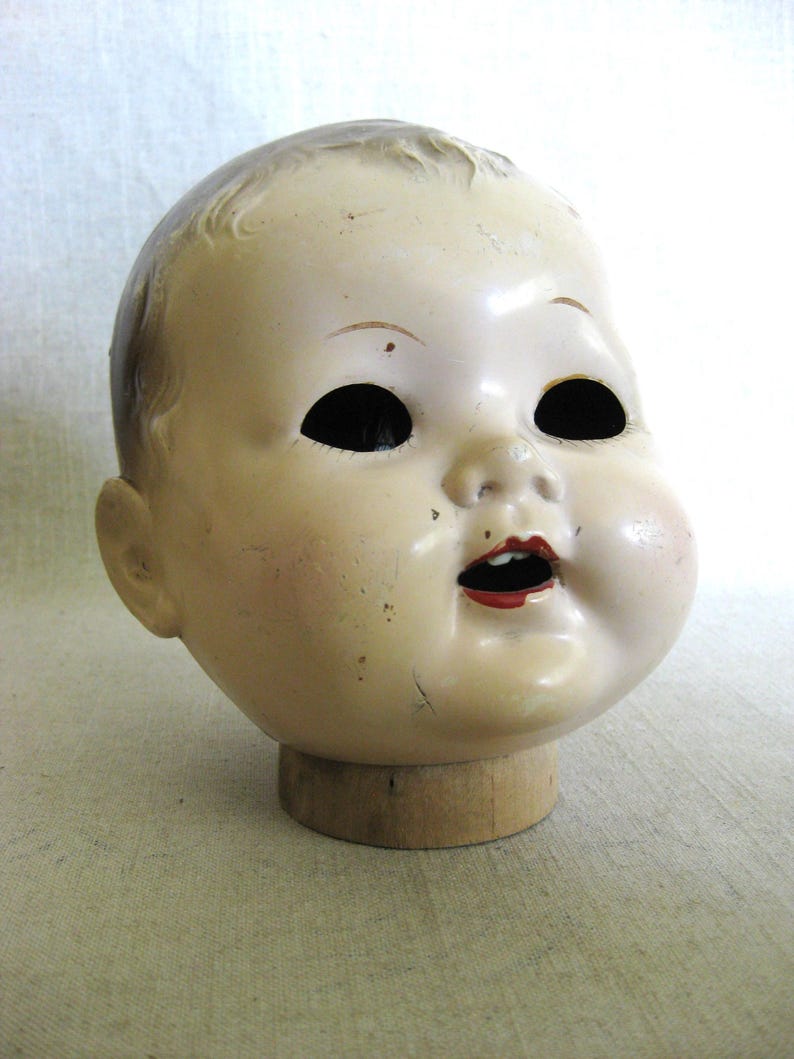 May include: Vintage doll head with a pale complexion, black eye sockets, and red lips. The doll head is mounted on a wooden base, showcasing its aged appearance and classic design. The head has a slightly open mouth.