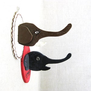 May include: Two wooden elephant head wall hooks, one brown and one black, mounted on a red oval plaque with white trim. The plaque has the word "Elephant" written on it.
