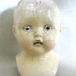 May include: A vintage doll head with a cracked, off-white finish. The doll head has black eye sockets and a small, closed mouth. The paint is peeling, revealing a network of cracks across the surface, giving it an antique appearance.
