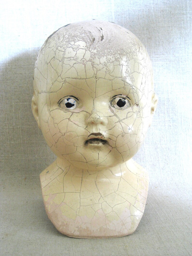 May include: A vintage doll head with a cracked, off-white finish. The doll head has black eye sockets and a small, closed mouth. The paint is peeling, revealing a network of cracks across the surface, giving it an antique appearance.