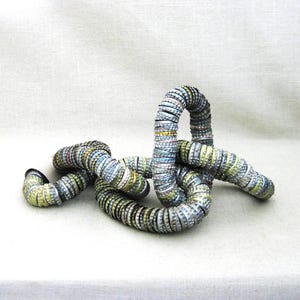 May include: Abstract sculpture crafted from repurposed bottle caps. The artwork features a looping, serpentine form with a mix of silver, yellow, and blue caps. The piece is set against a neutral background, highlighting its unique texture and design.