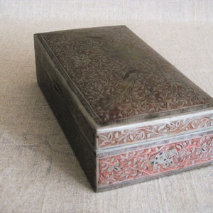 May include: A rectangular silver box with intricate floral designs engraved on the top and sides. The box has a red and silver border with additional floral designs.
