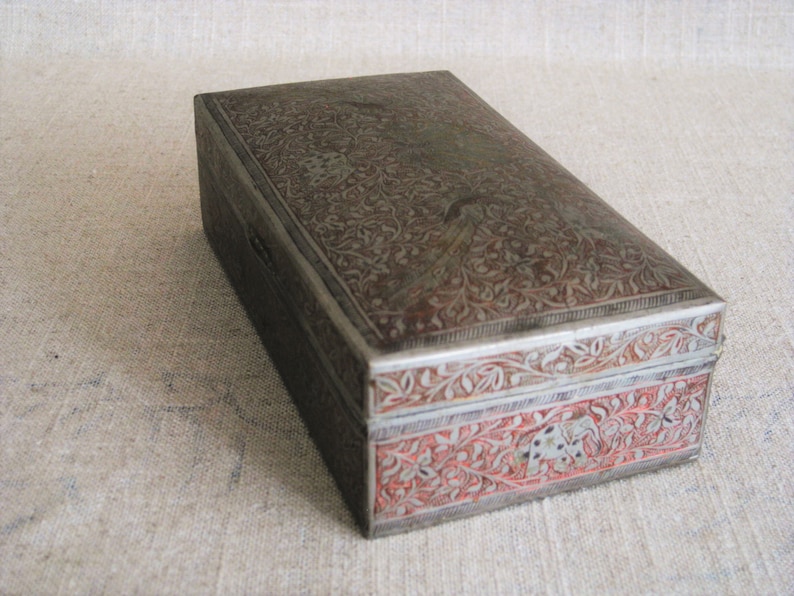 May include: A rectangular silver box with intricate floral designs engraved on the top and sides. The box has a red and silver border with additional floral designs.
