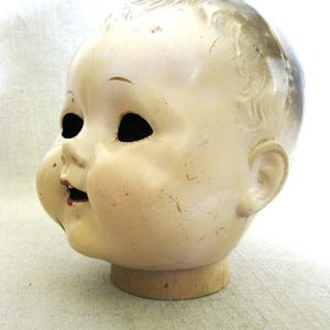 May include: Vintage doll head with a cream-colored face, black eye sockets, and a small red mouth. The head is mounted on a wooden base, showing signs of age and wear. The doll head has a slightly open mouth and a detailed, aged appearance.