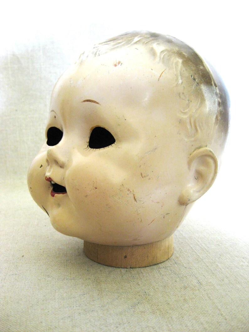 May include: Vintage doll head with a cream-colored face, black eye sockets, and a small red mouth. The head is mounted on a wooden base, showing signs of age and wear. The doll head has a slightly open mouth and a detailed, aged appearance.