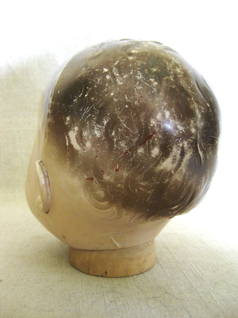 May include: Vintage doll head with painted features and brown hair. The head is mounted on a wooden base. The paint shows signs of wear, adding to its antique charm. The doll head is a collectible item.