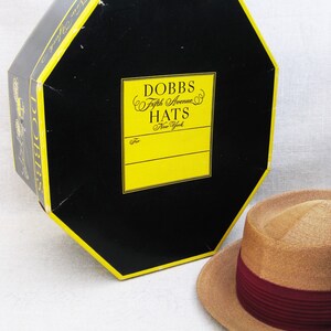 Pu&ograve; includere: Un cappello di paglia con una fascia bordeaux in una scatola che dice "Dobbs Fifth Avenue Hats New York".