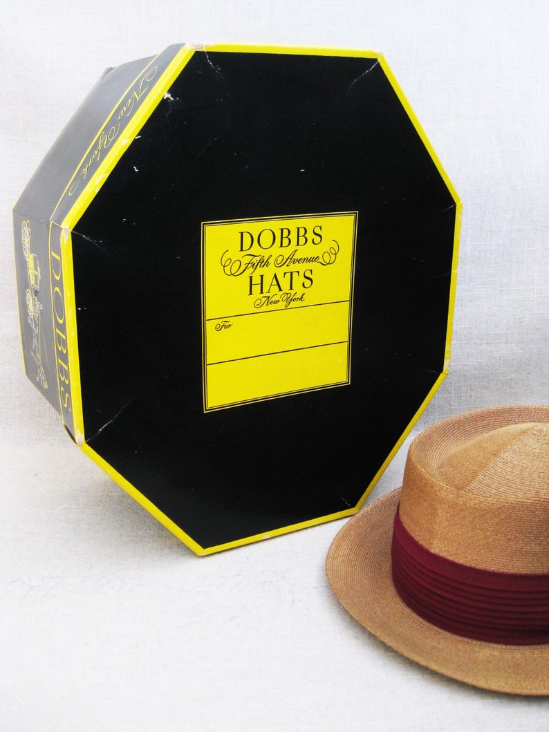 Pu&ograve; includere: Un cappello di paglia con una fascia bordeaux in una scatola che dice "Dobbs Fifth Avenue Hats New York".