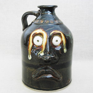 May include: A dark green ceramic jug with a face, white eyes, and a brown glaze. The jug has a handle and a spout.