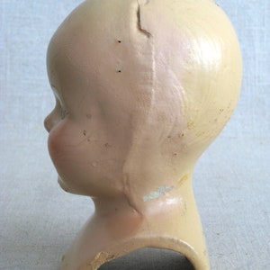 Puede incluir: Una cabeza de mu&ntilde;eca vintage hecha de yeso con una grieta en el centro. La cabeza est&aacute; pintada de un color rosa p&aacute;lido y tiene la boca ligeramente abierta. A la cabeza de la mu&ntilde;eca le faltan los ojos y el pelo.