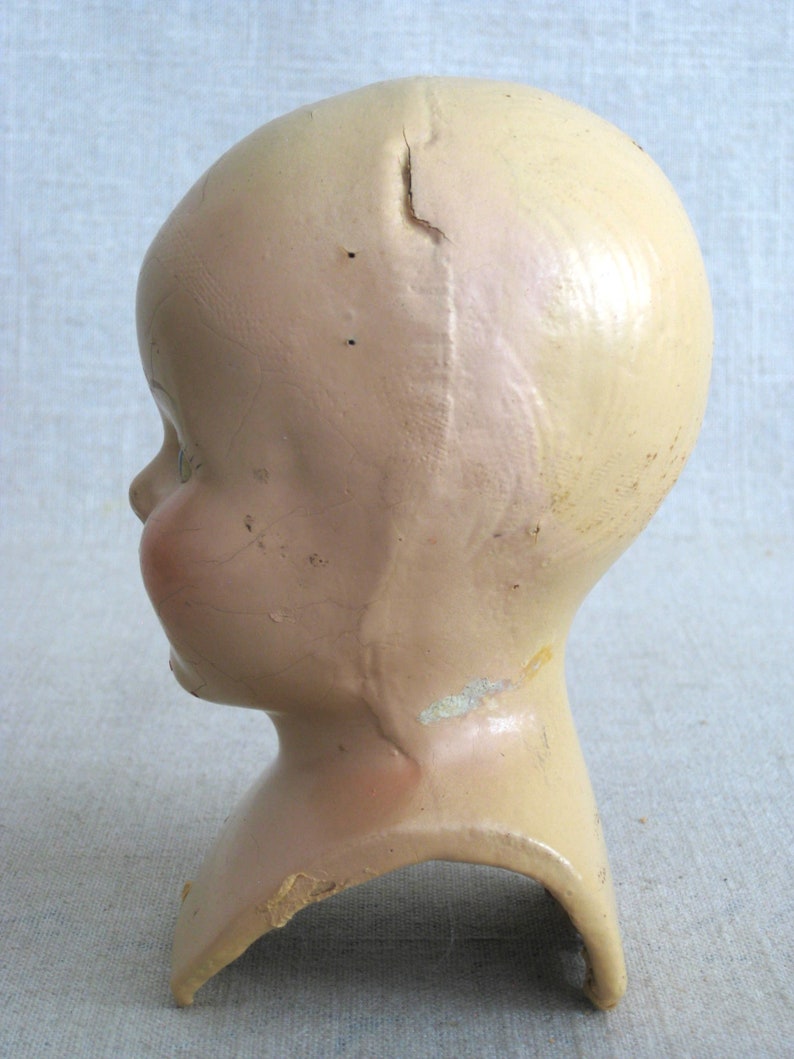 Puede incluir: Una cabeza de mu&ntilde;eca vintage hecha de yeso con una grieta en el centro. La cabeza est&aacute; pintada de un color rosa p&aacute;lido y tiene la boca ligeramente abierta. A la cabeza de la mu&ntilde;eca le faltan los ojos y el pelo.