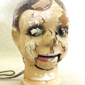 May include: An antique ventriloquist dummy head with a distressed, cracked paint finish. The head is painted in shades of beige, brown, and red, with visible wear and tear. A string is attached to the base. The mouth is open, revealing a red interior.