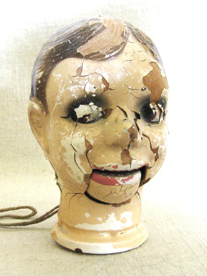 May include: An antique ventriloquist dummy head with a distressed, cracked paint finish. The head is painted in shades of beige, brown, and red, with visible wear and tear. A string is attached to the base. The mouth is open, revealing a red interior.