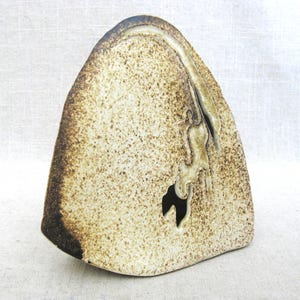 May include: A triangular ceramic sculpture with a speckled beige and cream surface. The piece features a dark brown, rough-textured edge and a central irregular cutout that reveals a dark interior space, creating a distinct organic form.