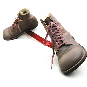 May include: Two brown leather boots with laces tied together with a red ribbon. The boots are on a white background.
