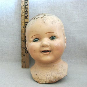 May include: An antique doll head with a serene expression and cracked porcelain. The doll head has blue-green eyes, rosy cheeks, and a slightly open mouth. A wooden ruler is placed next to the doll head.