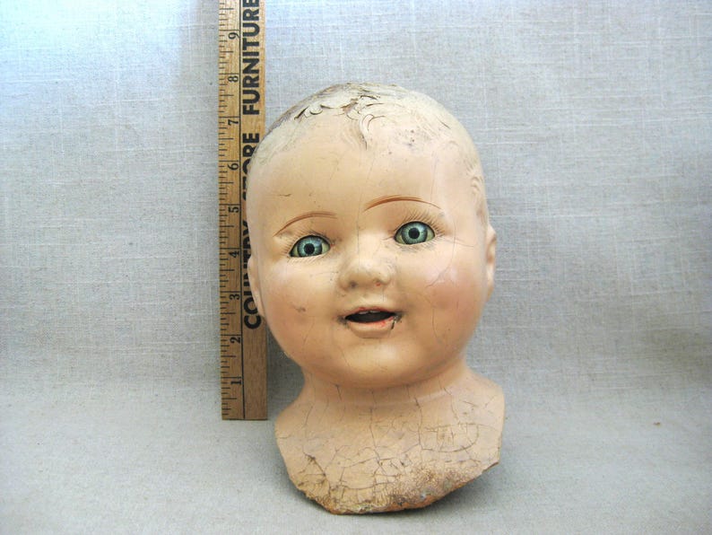 May include: An antique doll head with a serene expression and cracked porcelain. The doll head has blue-green eyes, rosy cheeks, and a slightly open mouth. A wooden ruler is placed next to the doll head.