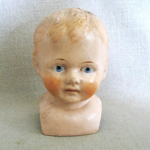 May include: Antique doll head with blue eyes and rosy cheeks. The doll head has blonde hair and a neutral skin tone. The doll head is a vintage item, likely porcelain or bisque.